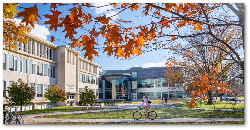 A single bicyclist rides by the Isenberg School of Management on a sunny day framed by orange fall leaves in the top foreground