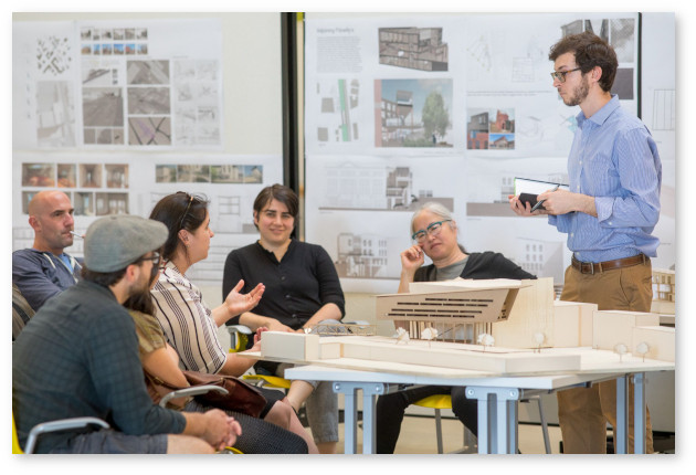 A small group of students and an instructor sit around a table discussing another, standing student's architecture assignment. There's a building model on the table, and the panels behind the presenting student are covered with elevtions and other buiding and site sketches.
