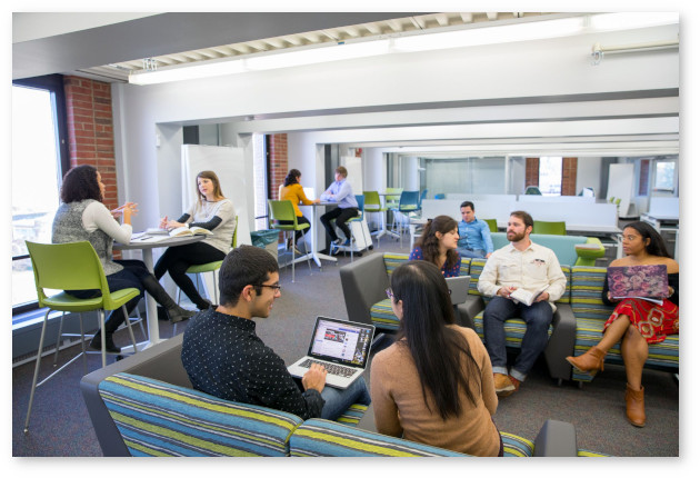 Students relax in Graduate Student Commons. Small groups of students sit at tables or on couches studying or conversing. The room is clean and contemporary with gray furniture with cornflower blue and chartreuse accents.