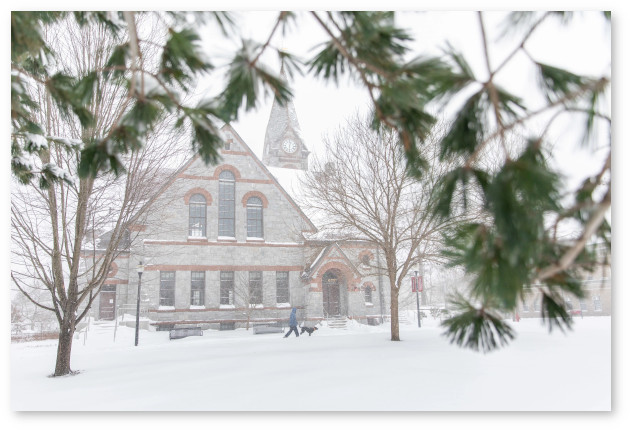Pine branches occupy the foreground of this picture of Old Chapel in the snow.  A person and their back dog walk past the building.