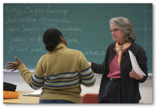 A young woman student facing away from the camera chats enthusasticaly with a woman professor, who faces us. Both are standing in front of a green chalkboard.