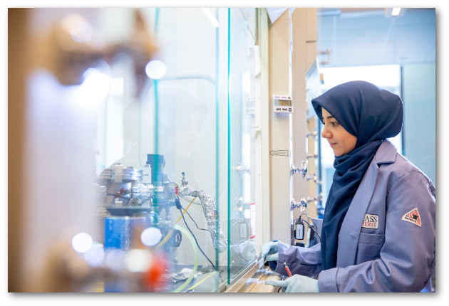 A young woman in a blue lab coat and head scarf monitors experimental equipment behind a glass panel.