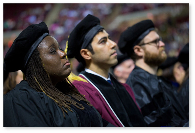Close up of three robed individuals sitting and facing to the right watching an unseen speaker during the graduate commencement ceremony.