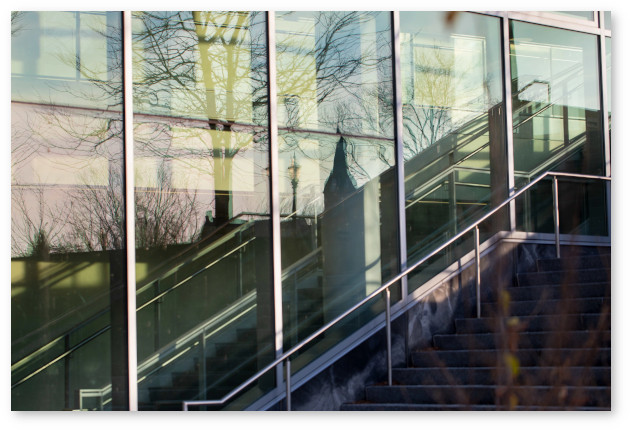 Tall exterior windows next to stairs outside the Integrative Learning Center reflect silhouettes of bare trees and the top of the Old Chapel spire.