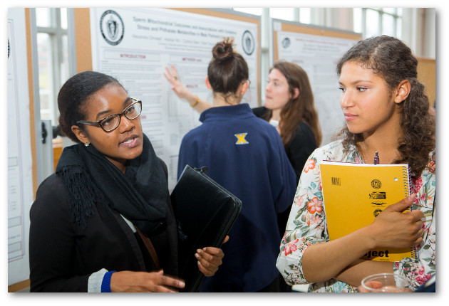 Two women undergraduates chat in foreground; poster session and additional women students in the background