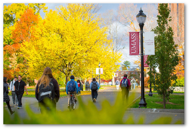 Students walking along campus mall in fall. UMass banners hang from lamp posts. Bright yellow and orange trees fill the left side of the image.