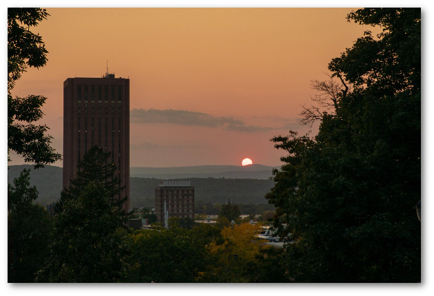 Evening view from Orchard Hill framed by trees looking down to Dubois Library and Thompson tower with sun setting behind Hadley hills