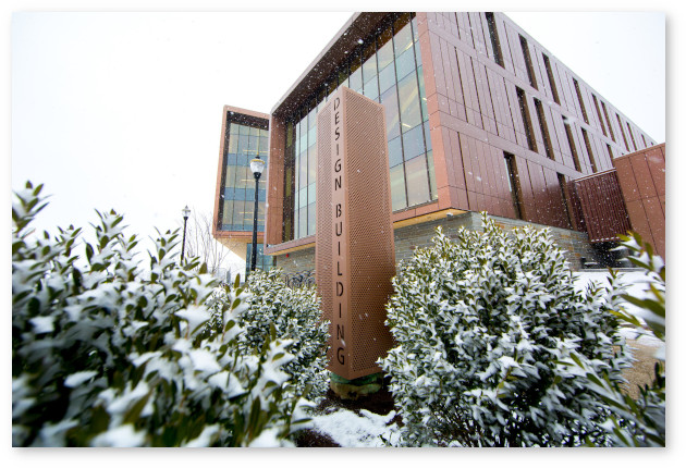 Looking up at the Olver Design Building against an overcast winter sky. Foreground bushes are dusted with snow.
