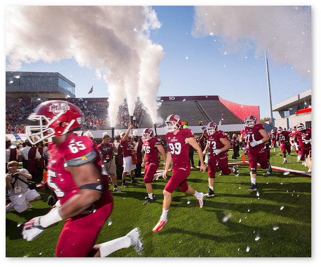 The UMass football team, wearing maroon uniforms, takes the field amid confetti and smoke canons during Homecoming.