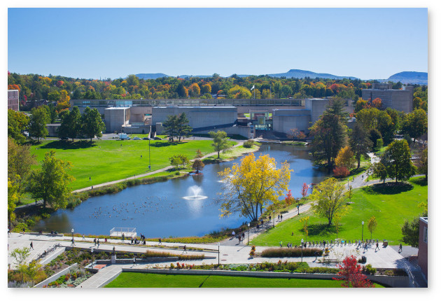 A summer view of the campus pond and Fine Arts Center from an upper library floor. The Holyoke range is visible in the background behind trees.