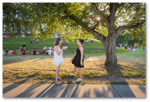 Two young women wearing casual summer dresses stand and chat face-to-face outside near a large tree. Groups of students sit on the grass in the background.