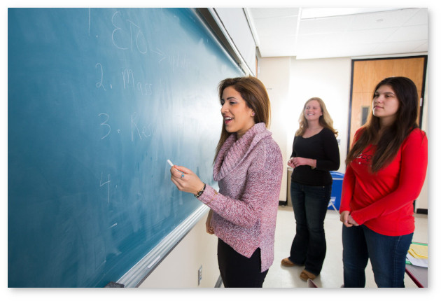 A teacher writes on a chalkboard while two students stand behind her and look on.