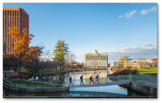 Four students crossing a narrow footbridge over one end of the geese-covered pond near the Fine Arts Center with Campus Center in the background