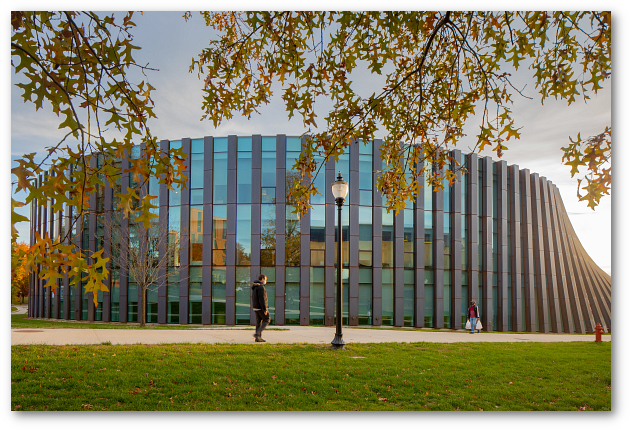 A single student walks by the new Isenberg addition, which features metallic vertical posts alternating with tall windows.  The wall changes from vertical to sloped as it winds around the curved building.