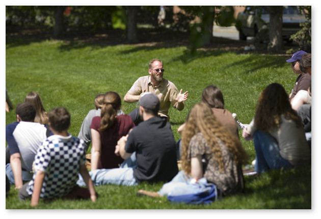 A group of students with their backs towards us sit on the grass and watch an animated instructor during an outdoor class.