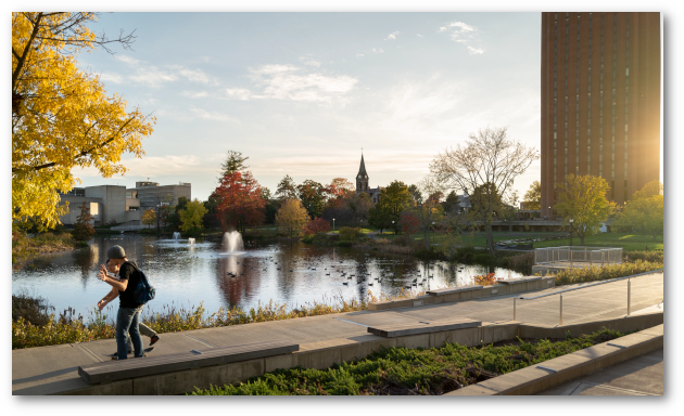 Two young men walk and skateboard along a sidewalk next to the campus pond at dusk.  Three fountains are spraying in the pond.  The library, Old Chapel. and Fine Arts Center are visible across the pond.