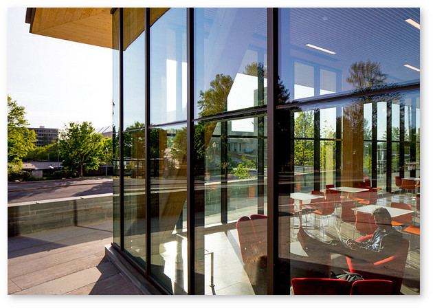 Looking into the Design Building lounge from outside, through the interior, then outside again to the Fine Arts Center.  A student working on a laptop has his feet up on one of the small tables visible in the glass-enclosed lounge.