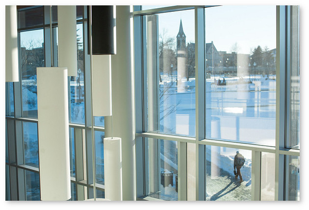 View through windows in the Integrated Sciences Building. Staggered vertical ight fixtures are shown in the foreground and reflected in the window. A snowy campus is visible outside.